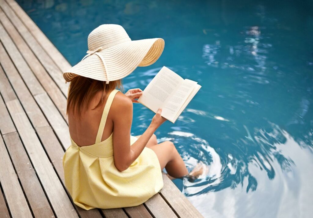 Frau im gelben Kleid und mit einem Hut liest ein Buch am Pool