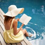Frau im gelben Kleid und mit einem Hut liest ein Buch am Pool