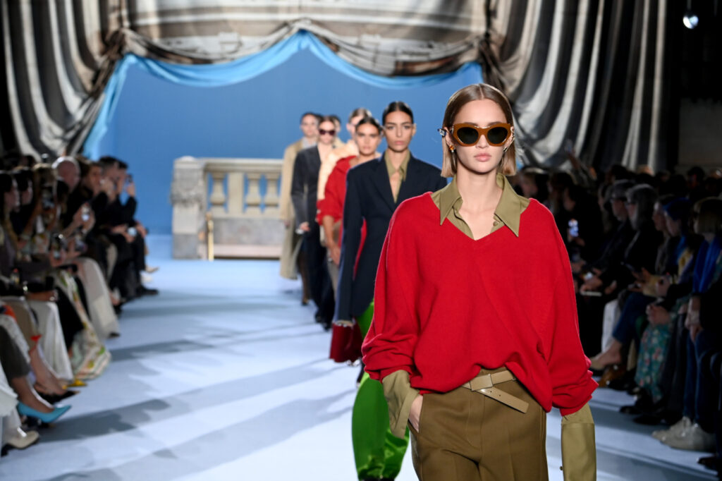 Foto von einer Modenschau, im Vordergrund ein Model in einem roten Pullover