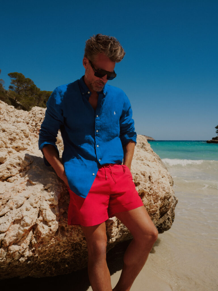 Mann am Strand in roten Badeshorts, blauem Hemd und Sonnenbrille